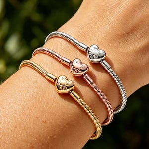 Op de afbeelding: Drie armbanden in goud, roségoud en zilver, elk met een hartvormige bedel. De armbanden worden om een pols gedragen, waarbij hun verschillende kleuren en ontwerpen worden getoond. De bedels hebben een gedetailleerd ontwerp.
