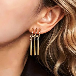 May include: Gold-colored hoop earrings with three dangling, elongated cone-shaped charms. The earrings are worn on a person's ear, with the focus on the jewelry's design and the warm tones of the gold against the skin and hair.