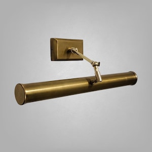 May include: A brass picture light fixture with a rectangular wall mount and an adjustable arm. The light has a cylindrical shade and a warm, metallic finish. The fixture is designed for illuminating artwork.