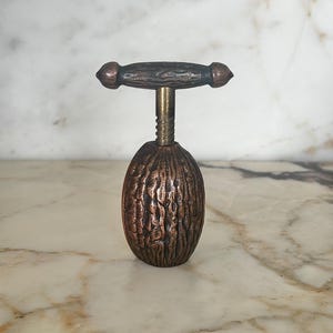 May include: A bronze-toned metal nutcracker featuring a textured, oval-shaped base resembling a walnut shell. The top section consists of a horizontal handle with a threaded screw mechanism that connects to the base, allowing for manual operation.