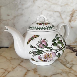 May include: A white ceramic teapot featuring a botanical illustration of pink flowers, green leaves, and a moth. The lid has a matching moth design and a green leaf border. The teapot includes a curved spout, a handle, and a circular base.