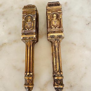 May include: Two matching gold-toned metal nutcrackers featuring an embossed portrait of a figure in historical attire within an arched frame. The handles are designed with vertical fluted columns and decorative bands, showing a weathered finish.