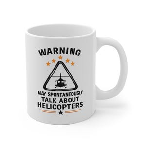 Könnte beinhalten: Weiße Keramik-Kaffeetasse mit dem Text "WARNING MAY SPONTANEOUSLY TALK ABOUT HELICOPTERS" in Schwarz und Orange. Die Tasse zeigt eine Hubschraubergrafik in einem Dreieck mit Sternen.
