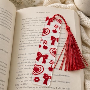 May include: A clear acrylic bookmark with a red tassel, featuring red bows, cherries, hearts, and flower designs. The bookmark is placed on an open book with the title "My Brother's Secret" visible.