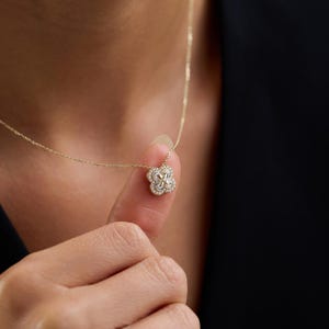 Könnte beinhalten: Eine zarte goldene Halskette mit einem vierblättrigen Kleeblatt-Anhänger. Der Anhänger ist mit kleinen, funkelnden Edelsteinen besetzt. Die Halskette wird von einem Finger gehalten, was das filigrane Design und die Handwerkskunst zeigt.