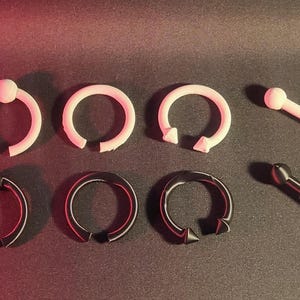 Puede incluir: Un surtido de joyas para el cuerpo, que incluye barras circulares rosas y negras, y barras rectas. Las joyas están dispuestas sobre una superficie oscura con un degradado de rojo a verde.