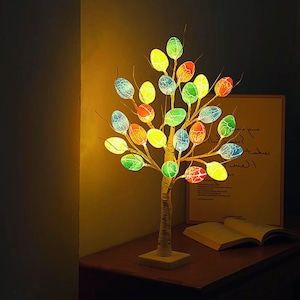 Puede incluir: Una lámpara decorativa con forma de árbol con luces coloridas en forma de huevo. El árbol tiene un tronco y ramas de color claro, con cada 'hoja' iluminada en tonos de amarillo, verde, naranja y azul. La lámpara se apoya en una base de madera.