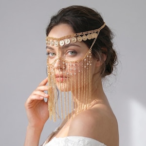 May include: A gold-toned headpiece featuring a delicate chain design that drapes across the forehead and cheeks. The accessory is adorned with small, circular metallic discs and fine hanging chains that create a layered, fringe-like effect.