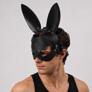 Op de afbeelding: Zwart leren konijnenmasker met lange, rechtopstaande oren. Het masker bedekt de ogen en neus, vastgezet met een band om het hoofd. Klinknagels en gebogen lijnen defini&euml;ren het ontwerp. De persoon draagt een zwarte tanktop.