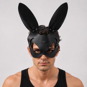Op de afbeelding: Zwart leren konijnenmasker met lange, rechtopstaande oren. Het masker bedekt de ogen en neus, vastgezet met klinknagels en riemen. De persoon op de foto draagt een zwarte tanktop.
