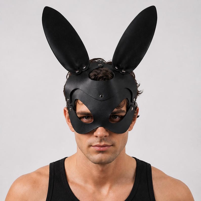 Op de afbeelding: Zwart leren konijnenmasker met lange, rechtopstaande oren. Het masker bedekt de ogen en neus, vastgezet met klinknagels en riemen. De persoon op de foto draagt een zwarte tanktop.