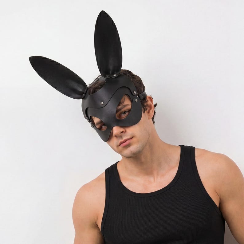 Op de afbeelding: Een zwart leren konijnenmasker met lange, rechtopstaande oren. Het masker bedekt de ogen en neus, vastgezet met riemen en klinknagels. De persoon draagt een zwarte tanktop.