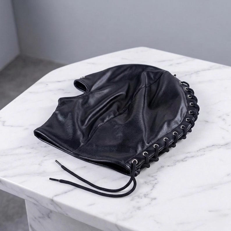 Puede incluir: Capucha de cuero negro con dise&ntilde;o de cordones en el lateral. La capucha tiene una textura suave y est&aacute; atada con cordones negros. El producto se exhibe sobre una superficie de m&aacute;rmol blanco.