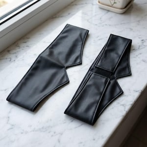 Op de afbeelding: Twee zwarte hoofdbanden van kunstleer met een puntig, hoekig ontwerp. Elke hoofdband heeft een gladde, matte afwerking en een gestructureerde vorm die naar het midden toe taps toeloopt. Eén hoofdband toont een zichtbare zwarte klittenbandsluiting.