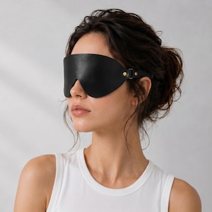Könnte beinhalten: Eine schwarze Augenbinde aus Leder mit einer glatten, konturierten Form, die die Augen bedeckt. Die Augenbinde wird mit einem dünnen Riemen und kleinen goldfarbenen Metallbeschlägen, einschließlich Nieten und einer Schnalle, an der Seite befestigt.