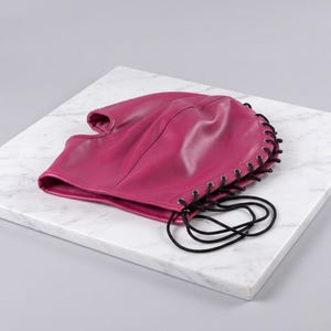 Puede incluir: Una capucha de cuero magenta con una parte superior redondeada y una sola abertura para la cara. Los cordones negros recorren la parte posterior y los lados, formando una atadura. La capucha est&aacute; sobre una superficie de m&aacute;rmol blanco.