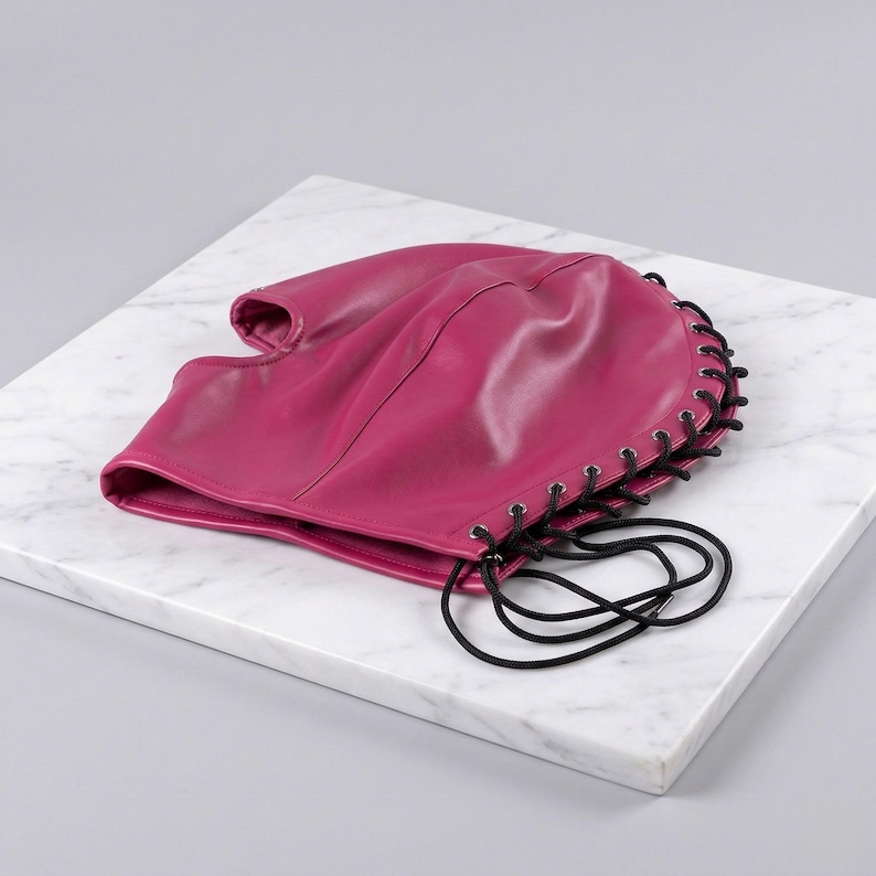 Puede incluir: Una capucha de cuero magenta con una parte superior redondeada y una sola abertura para la cara. Los cordones negros recorren la parte posterior y los lados, formando una atadura. La capucha est&aacute; sobre una superficie de m&aacute;rmol blanco.