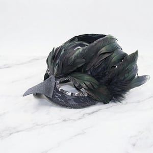 May include: A black masquerade mask with dark feathers, a grey beak, and a decorative lace and sequin trim. The mask is designed for a formal event or costume party.