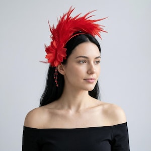 May include: A vibrant red feather headband with a large flower and beaded accents. The headband features a cluster of feathers and a fabric flower on the side. The accessory is designed to be worn on the head.