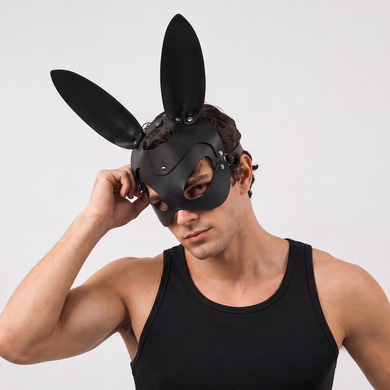 Op de afbeelding: Zwart leren konijnenmasker met lange, rechtopstaande oren. Het masker bedekt de ogen en neus, vastgezet met riemen en klinknagels. De persoon op de foto draagt een zwarte tanktop.