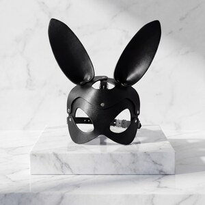 Op de afbeelding: Zwart leren konijnenmasker met lange, rechtopstaande oren. Het masker heeft ooggaten en een riem met een gesp voor een veilige pasvorm. Het masker is ontworpen met klinknagels en wordt gepresenteerd op een wit marmeren oppervlak.