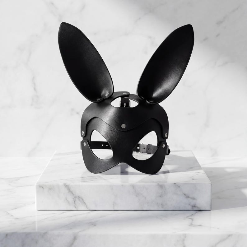 Op de afbeelding: Zwart leren konijnenmasker met lange, rechtopstaande oren. Het masker heeft ooggaten en een riem met een gesp voor een veilige pasvorm. Het masker is ontworpen met klinknagels en wordt gepresenteerd op een wit marmeren oppervlak.