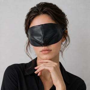 May include: A black faux leather sleep mask with a smooth, matte finish and a central vertical seam. The mask features a contoured shape designed to cover the eyes completely, with a flexible strap attached to the sides for a secure fit.