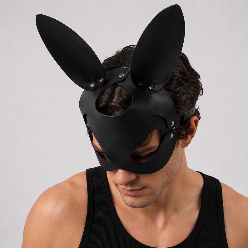 Op de afbeelding: Zwart leren konijnenmasker met lange, rechtopstaande oren. Het masker heeft uitsparingen voor ogen en neus, vastgezet met klinknagels en een riem. Het masker is ontworpen om het gezicht te bedekken.