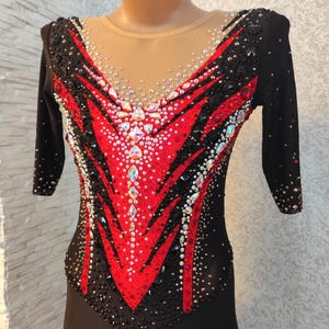 May include: Black and red leotard with jewel embellishments. The leotard features a red and black design on the bodice, with a sheer neckline and long sleeves. Rhinestones and crystals are scattered across the front, adding sparkle and dimension to the garment.