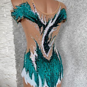 May include: A teal and nude leotard with a decorative design. The leotard features a teal bodice with a white skirt and is embellished with rhinestones. The design includes a pattern of dark green and white accents.