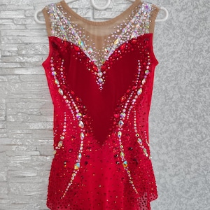 May include: A red sleeveless leotard with a heart-shaped velvet panel on the bodice. The leotard is adorned with rhinestones and crystals, and features a nude mesh neckline. The garment is designed for performance.