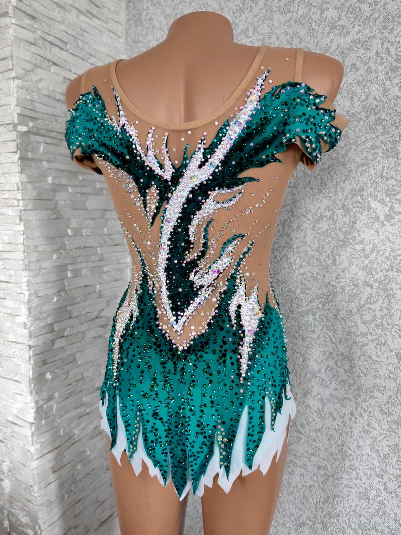 May include: A teal and nude-coloured leotard with a decorative design. The leotard features a teal bodice with white accents and is embellished with sparkling rhinestones. The design extends up the back and over the shoulders.
