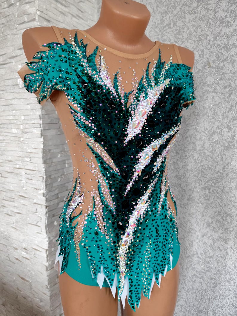 May include: A teal and black leotard with a nude mesh overlay, adorned with sparkling rhinestones. The leotard showcases a distinctive feather-like design, with a high neckline and short sleeves. The bottom hem has a white, jagged edge.