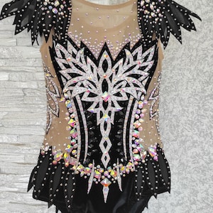 May include: A black and nude leotard with crystal and sequin detailing. The bodice has a large, symmetrical design with a central floral motif. Black mesh wings extend from the shoulders, adding a dramatic flair to this performance garment.