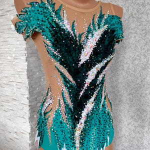 May include: A teal and black leotard with a nude mesh overlay, adorned with sparkling rhinestones. The design features a feather-like pattern across the bodice and sleeves, with a teal skirt and white accents. This garment is likely for gymnastics or dance.