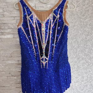 May include: A blue sleeveless leotard with detailed sequin and rhinestone embellishments. The bodice has a tan mesh neckline and a pattern of vertical lines. The skirt is pleated. The garment is displayed on a white hanger.