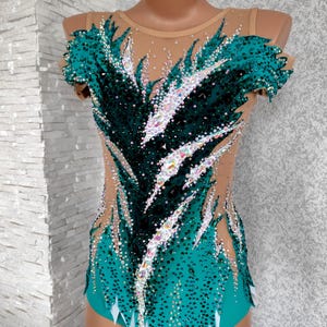 May include: A teal and black leotard with a nude mesh overlay, adorned with sparkling rhinestones. The design features a feather-like pattern across the bodice and sleeves, with white accents at the hem. Suitable for gymnastics or dance performances.