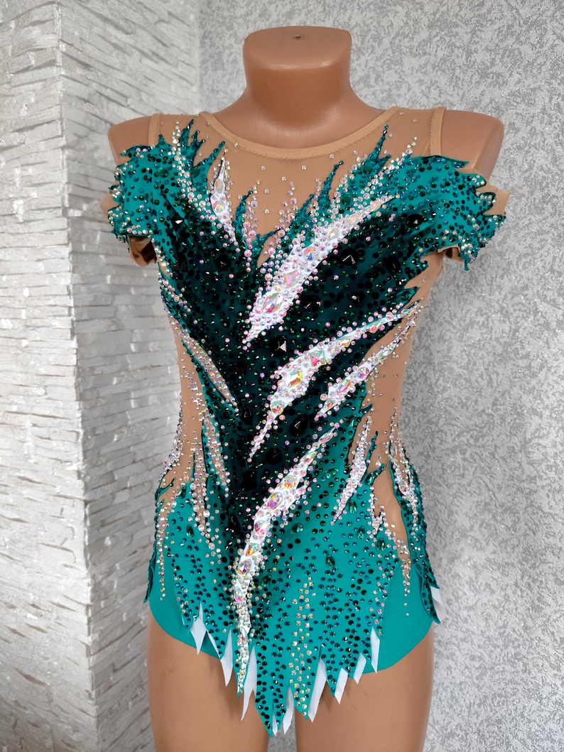 May include: A teal and black leotard with a nude mesh overlay, adorned with sparkling rhinestones. The design features a feather-like pattern across the bodice and sleeves, with white accents at the hem. Suitable for gymnastics or dance performances.