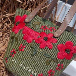 Peut inclure: Sac fourre-tout en raphia vert avec des fleurs rouges brodées et des anses en cuir marron. Le sac porte le texte "st raffia" et présente une texture tissée. Les anses sont fixées avec des rivets en laiton.