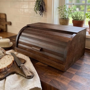 May include: A dark brown wooden bread bin with a roll-top lid sits on a butcher block countertop. Sliced bread and a bread knife rest on a wooden chopping board. A butter dish and herbs are also visible.