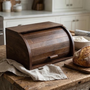 Puede incluir: Una panera de madera marrón oscuro con tapa enrollable y un pequeño asa. La panera está sobre una mesa de madera junto a una hogaza de pan y un paño de lino. Ambiente de cocina.