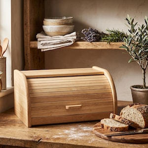 Op de afbeelding: Een houten broodtrommel met een rolluikdeksel en een klein handvat. Gesneden brood op een houten snijplank ernaast. Op de achtergrond planken met kommen, kruiden en een kleine potplant. De broodtrommel staat op een houten oppervlak.