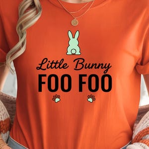 May include: Orange t-shirt with the text "Little Bunny Foo Foo" in black script. A light green bunny graphic is above the text, with paw prints below. The shirt has rolled-up sleeves.