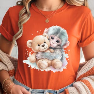 May include: Orange t-shirt featuring a watercolor illustration of a gray monkey hugging a tan teddy bear. The design is centered on the shirt. The shirt has rolled-up sleeves and is being worn by a person.