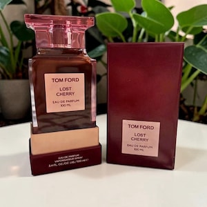 May include: A Tom Ford "Lost Cherry" eau de parfum bottle and box. The perfume bottle is rectangular with a pink cap and a brown liquid. The box is a deep red color with the perfume name and brand.