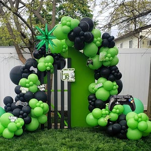 May include: A green and black balloon arch and decorations for a video game-themed birthday party. The decorations include balloons, signs with text like "Level Up" and "Game On! Birthday", and a controller-shaped balloon.