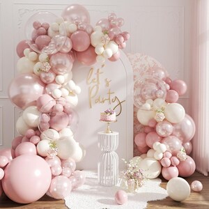 May include: A party scene with a balloon arch in shades of pink and white, with a small cake on a pedestal. The words "let's Party" are in gold script. The scene is decorated with flowers.