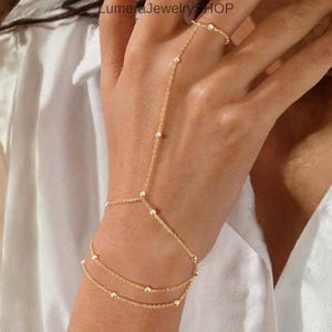 Puede incluir: Una delicada cadena de mano dorada con un diseño de anillo, muñeca y cadena de dedo. La joyería presenta pequeñas cuentas espaciadas uniformemente a lo largo de las cadenas. La cadena de mano se usa en la mano y la muñeca de una persona.