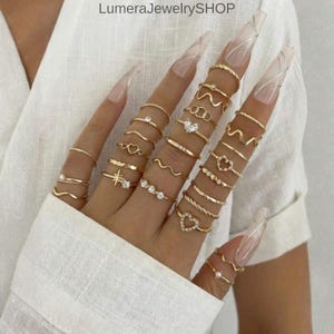May include: A selection of gold rings in diverse designs, including bands, hearts, and wave patterns. Some rings feature small clear gemstones. The rings are displayed on a hand, showcasing the different styles and sizes.