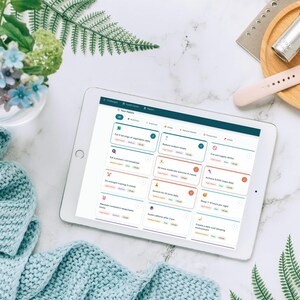 May include: A white tablet showing a health app interface, alongside a light blue knitted scarf, flowers, a wooden tray with a smartwatch, and other items on a white marble surface. The app interface displays health tips and advice.